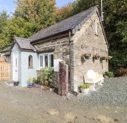 A stone cottage with a white door and window surrounded by plants and a bench outside at Pendre Cottage in Cenarth