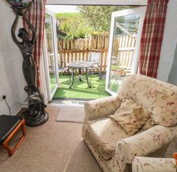 A sitting area with an armchair and a small table lamp next to open doors leading to a patio with a table and chairs at Pendre Cottage in Cenarth