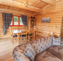 A wooden dining table with chairs near a window with curtains and a sofa with cushions in a log cabin living room at Maple Lodge in Greetham Rutland