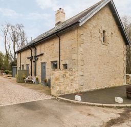 Exterior view of a stone building with multiple doors and benches near a gravel and paved driveway at Grange Cottages in Alnwick
