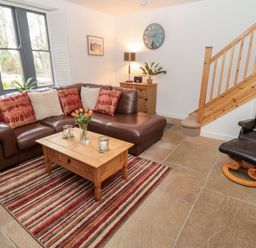 A living room with a brown leather sectional sofa red and beige cushions a wooden coffee table striped rug a wooden sideboard with a lamp and clock and a leather armchair with a deer cushion next to wooden stairs at Grange Cottages in Alnwick