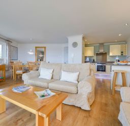 A living room with beige sofas a wooden coffee table dining table kitchen area and large window at 17 Burgh Island Causeway in Bigbury-On-Sea