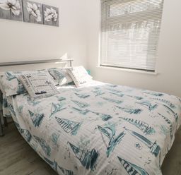 A bedroom with a bed with boat-themed bedding a window with blinds two side tables with blue lamps and wall art with flowers at Ballasalla in Liskeard