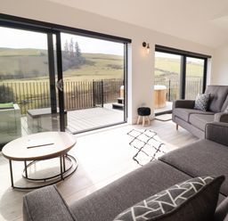 A living room with gray sofas, patterned cushions, a wood table, a small black stool, and glass sliding doors opening to a deck with a hot tub and a scenic countryside view at The Sheep Shed in Llanrhaeadr-Ym-Mochnant