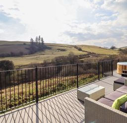 An outdoor deck with patio furniture a glass table and a hot tub overlooking grassy hills and trees at The Sheep Shed in Llanrhaeadr-Ym-Mochnant