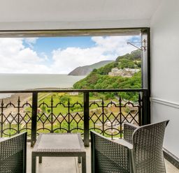 A balcony with two chairs and a table overlooking a coastal landscape with greenery and buildings at Clooneavin Apartment 4 in Lynmouth