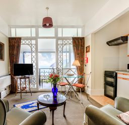 A living room with armchairs a small round table and a vase of flowers a TV on a stand and a glass dining table with chairs next to a kitchen area with appliances and cabinets at Clooneavin Apartment 4 in Lynmouth