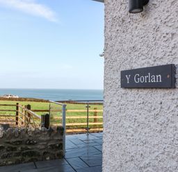 A view of a stone wall and outdoor area with a sign reading Y Gorlan and a grassy field and ocean in the background at Y Gorlan in Moelfre