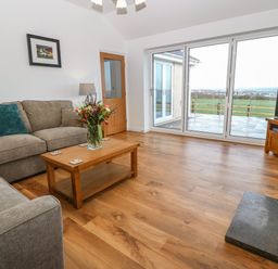 A living room with two sofas a wooden coffee table with flowers large sliding glass doors and a television at Y Gorlan in Moelfre
