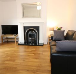 A living room with a black leather sofa a fireplace a TV on a wooden stand and a side table with a lamp and flowers at Gatehouse in Moville County Donegal