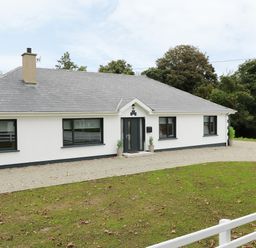 A single story house with a gray roof white walls and black window frames surrounded by grass and trees at Gatehouse in Moville County Donegal