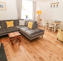 A living room with a gray sectional sofa yellow and white cushions a wooden table a basket of wood a dining table with chairs and a yellow armchair at Curlew Cottage 2 Fenkle Street Holy Island