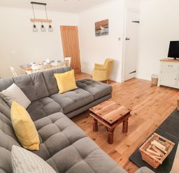 A living room with a gray sectional sofa yellow cushions wooden coffee table dining table with white chairs and a television on a cabinet at Curlew Cottage 2 Fenkle Street Holy Island