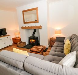 A living room with a gray sectional sofa yellow and patterned pillows two side tables with lamps a wood stove with fire a mirror above and a TV on a white cabinet at Curlew Cottage 2 Fenkle Street Holy Island