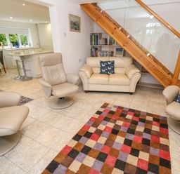 A living room with beige armchairs and sofa with patterned cushions a colorful checkered rug and a bookshelf under a wooden staircase at Alice Cottage in Wheal Kitty near St Agnes