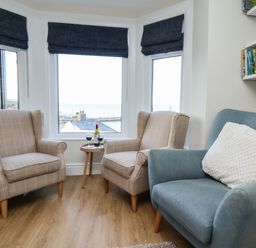 A sitting area with two beige armchairs and a blue chair with a white cushion by three windows with dark blinds and a small round table with wine glasses and a bottle at Mariners Watch in Whitby