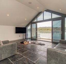 A living room with gray sofas a round coffee table a wall-mounted TV and large windows overlooking a lake at Boathouse on the Lake in Bowness-On-Windermere