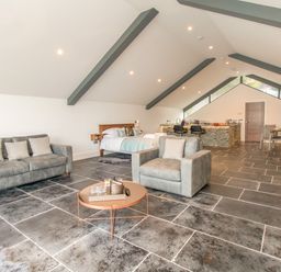 An open-plan room with a gray sofa and armchair around a round coffee table a bed near the back and a kitchen with bar stools and a dining area at Boathouse on the Lake in Bowness-On-Windermere
