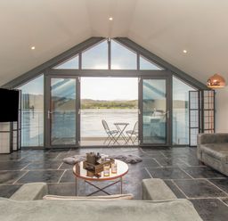 A living room with sofas a round coffee table and large glass doors opening to a lake view at Boathouse on the Lake in Bowness-On-Windermere