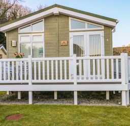 A small wooden cabin with a white railing and a patio table with flowers on the porch at The Hideaway - Heritage Park in Stepaside near Kilgetty