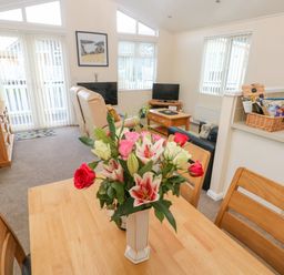 A dining table with a vase of flowers in a living room with sofas televisions and a cabinet at The Hideaway - Heritage Park in Stepaside near Kilgetty