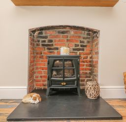 A black stove in a brick fireplace with a candle on top a cat curled up on the hearth and a wicker lantern beside it at Seasides in Weymouth