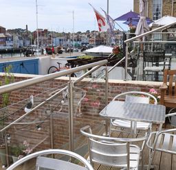 An outdoor seating area with metal chairs and table wooden bench glass railing overlooking a harbor with boats and buildings at Seasides in Weymouth