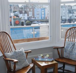 Two wooden chairs with cushions and a small wooden table with drinks in front of large windows overlooking a waterfront at Seasides Weymouth