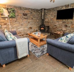 A living room with stone walls two blue sofas a wooden coffee table a wood stove and a wall-mounted TV at Granary Cottage in Bellingham