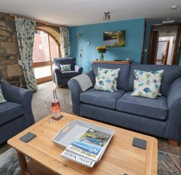 A living room with blue sofas floral cushions a wooden table with a bottle and glasses and stone walls at Granary Cottage in Bellingham