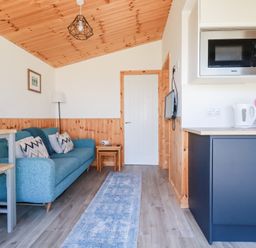 A living area with a blue sofa and cushions next to a wooden table and stools a kitchen counter with a microwave kettle and a pink tray with a crab design and a television mounted on the wall at 194 Atlantic Bays in Atlantic Bays Holiday Park near St Merryn