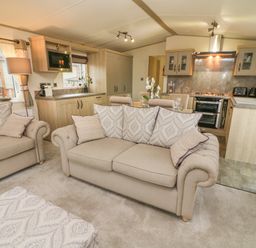A living room with beige sofas and a patterned ottoman next to a kitchen with wooden cabinets at Lancaster Lodge in South Lakeland Leisure Village