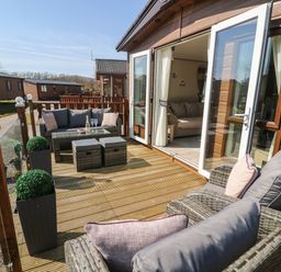 A wooden deck with wicker furniture and cushions outside a lodge with an open door at Lancaster Lodge South Lakeland Leisure Village
