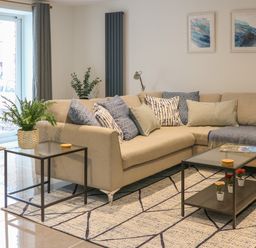 A living room with a beige sectional sofa with patterned cushions a glass coffee table with small potted flowers a side table with a plant sliding glass doors and two framed artworks on the wall at Fig Cottage in Kirkby Stephen