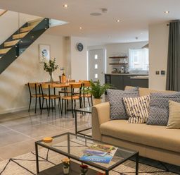An open living and dining area with a beige sofa with patterned cushions a glass coffee table with books a wooden dining table with chairs and a staircase with glass railing at Fig Cottage in Kirkby Stephen