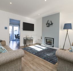 A living room with two sofas cushions a fireplace floor lamp a wall mounted tv and a dining area visible through a doorway at Beach Cottage in Portreath