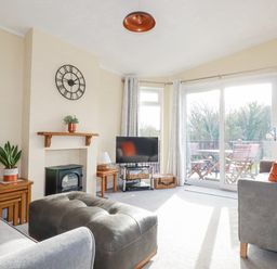 A living room with a clock above a fireplace a television and sofas with cushions at Laurel Lodge Bossiney Bay Holiday Park near Tintagel