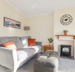 A living room with a grey sofa, orange pillows, a black stove, a wall clock, and wooden side tables at Laurel Lodge Bossiney Bay Holiday Park near Tintagel