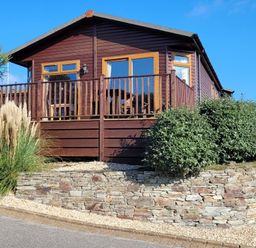 A wooden cabin with a balcony surrounded by bushes and ornamental grass at Laurel Lodge Bossiney Bay Holiday Park near Tintagel