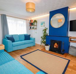 A living room with two blue sofas a wooden floor a fireplace with a round mirror above a mounted television and shelves with plants and books at Estuary View in Newquay