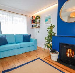 A living room with a blue sofa two cushions a burning fireplace round mirror plants and wooden shelves at Estuary View in Newquay