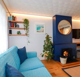 A living room with a blue sofa and cushions a wood burning stove with a round mirror above and shelves with books and plants at Estuary View in Newquay