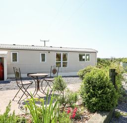 A metal table with two chairs on a patio next to a beige mobile home with propane tanks and greenery at The Lodge in Pontyates