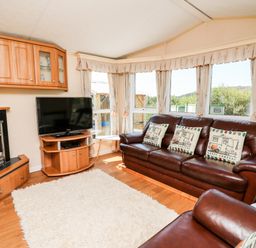 A living room with leather sofas, cushions, a television on a wooden stand, a fireplace, large windows with curtains, and a white rug on the wooden floor at The Lodge in Pontyates