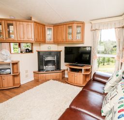 A living room with a brown leather sofa, a television on a wooden stand, a fireplace, wooden cabinets, and large windows with curtains at The Lodge in Pontyates