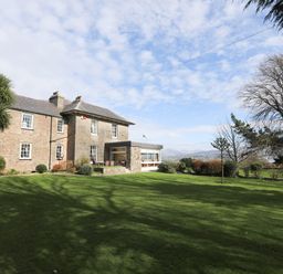 A large stone building with multiple windows surrounded by a lawn and trees at Tal Gwynedd in Dwyran near Newborough