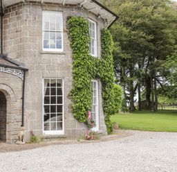 The exterior of a stone house with ivy on the walls two dog statues near an arched doorway and a lawn with trees at Tal Gwynedd in Dwyran near Newborough