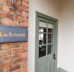 A green door with glass panes next to a brick wall with a Tackroom sign at The Tack Room in Lydney