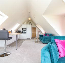 An open plan room with a kitchen area including stools and appliances and a sitting area with teal sofas and pink cushions at Higher Kernick View in Helland near Blisland
