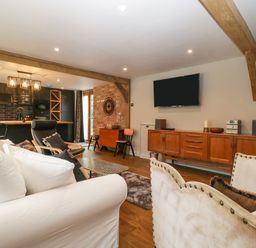 An open plan living room and kitchen with wooden beams a white sofa a black armchair and a wooden sideboard with a wall mounted TV at The Courtyard - Hilltop Barn in Winterborne Zelston near Wareham